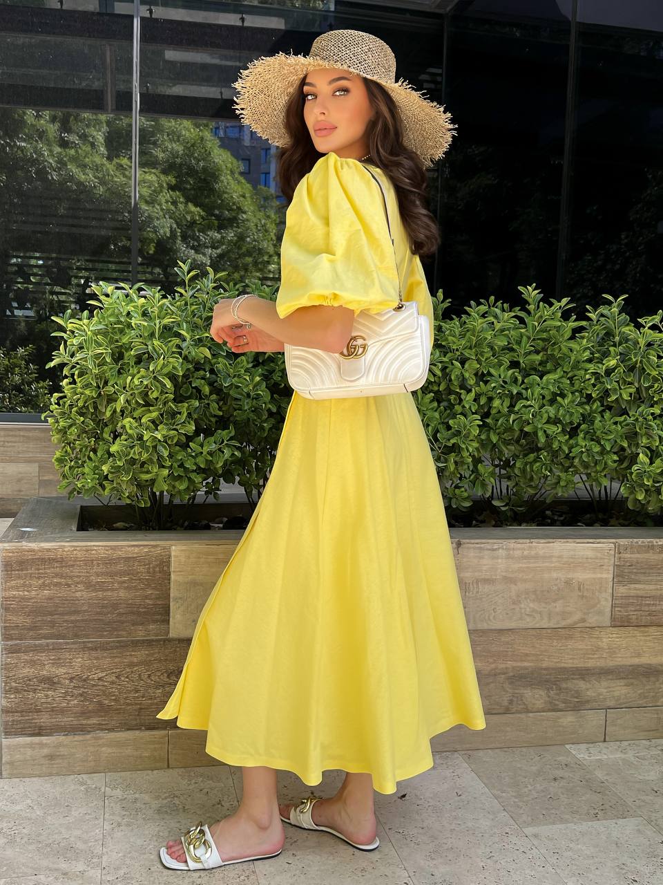 Woman in a yellow dress and hat standing outdoors with greenery in the background