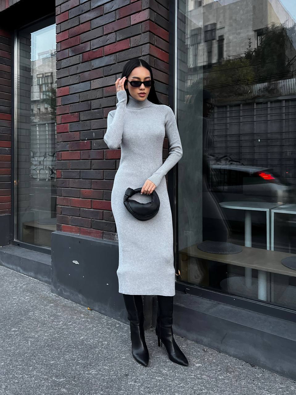 Woman in a gray dress and black boots standing against a brick wall.