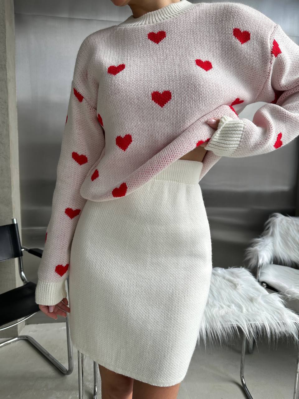 Person wearing a pink sweater with red heart patterns and a white skirt indoors.