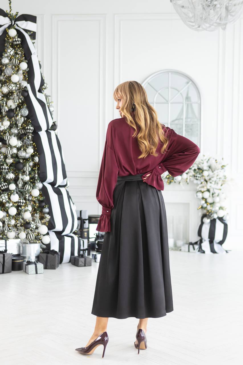 Woman in a burgundy blouse and black skirt standing in a decorated room with Christmas trees and presents.