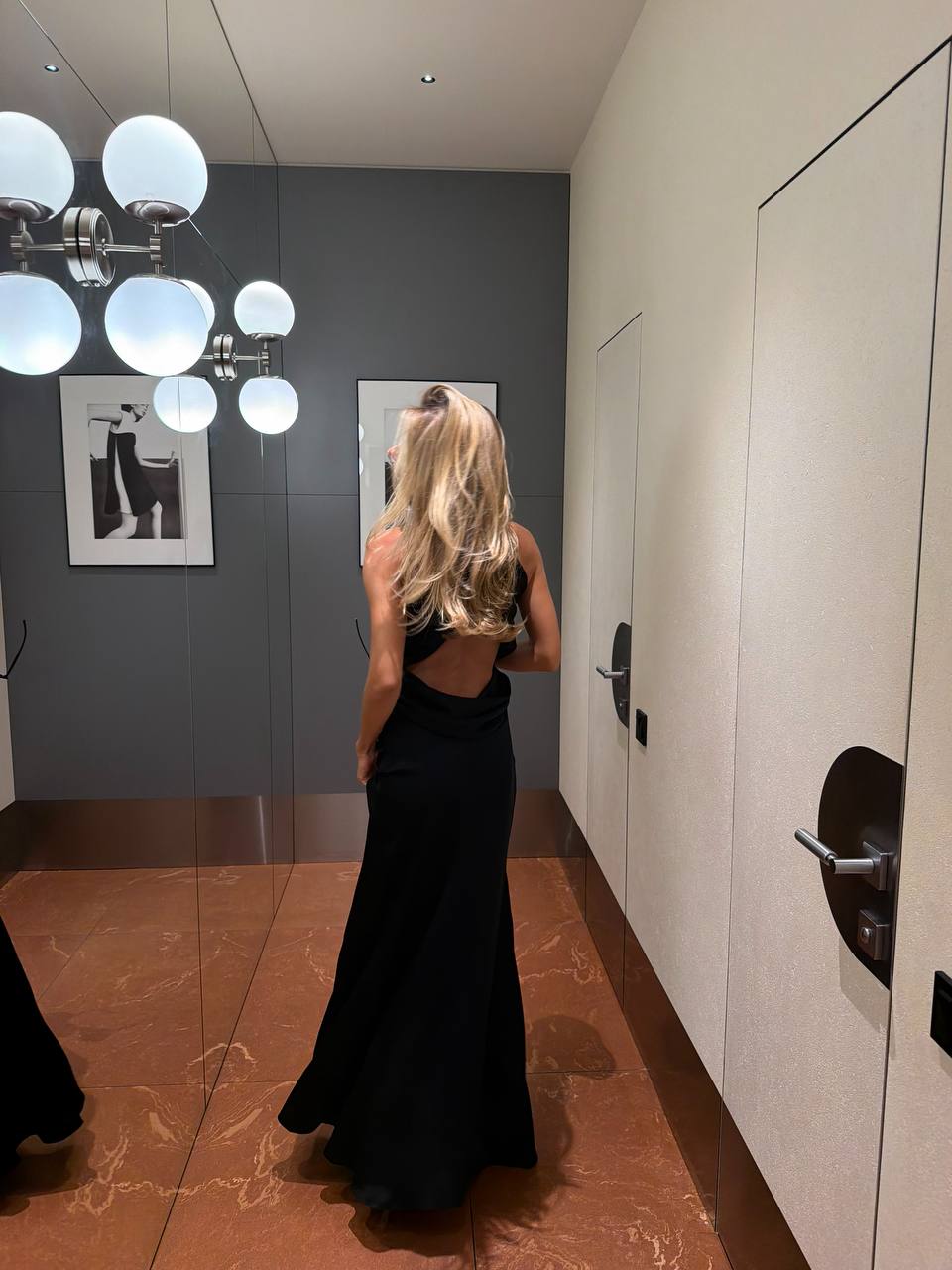 A woman with blonde hair stands with her back to the camera wearing a black backless evening gown in a modern hallway with a mirror, modern chandelier, and doors.