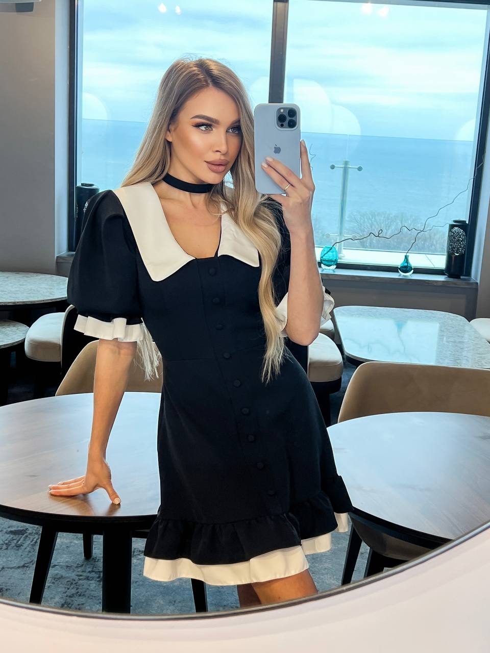 Woman taking a mirror selfie wearing a black dress with white collar and cuffs in an indoor setting.