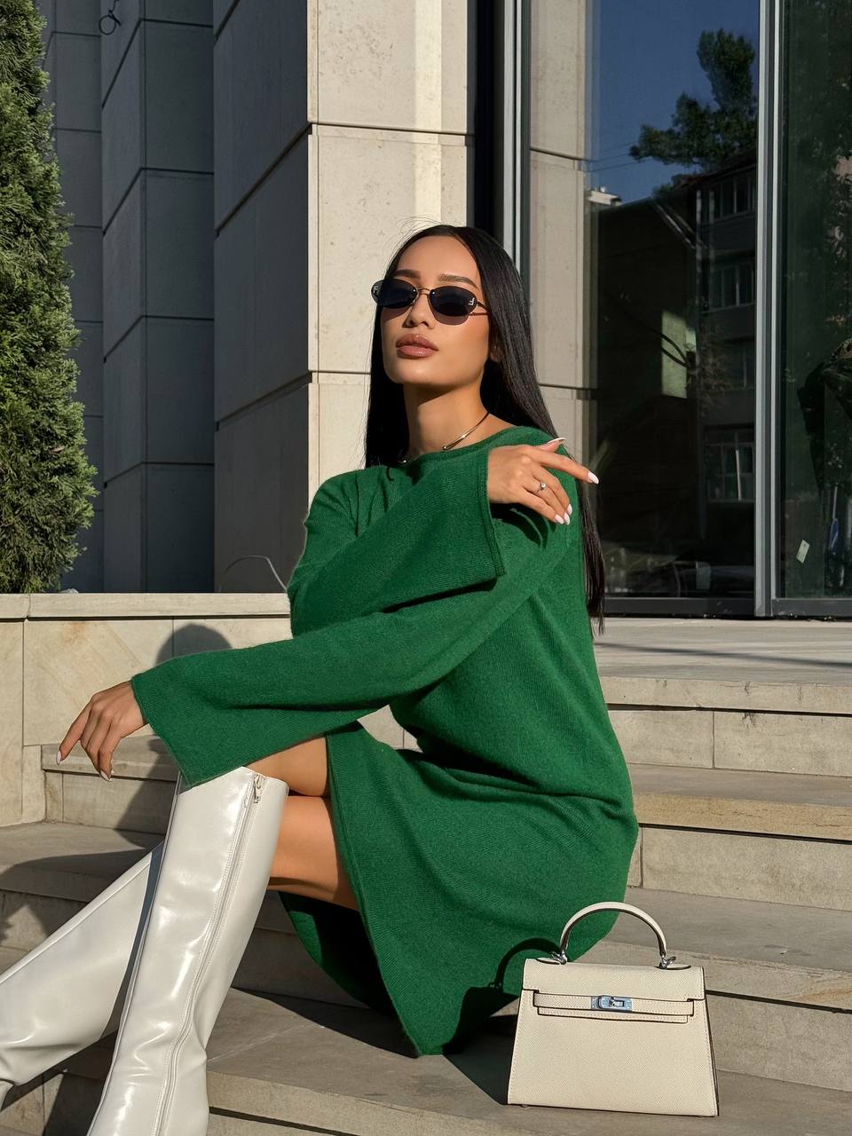 Woman in a green dress and white boots sitting on steps with a white handbag.