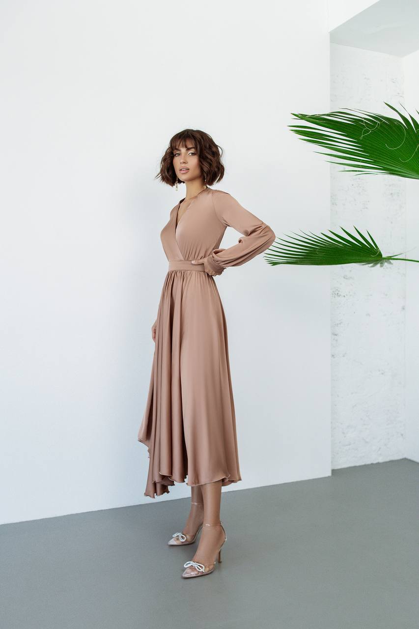 Woman wearing a beige dress standing against a white wall with a plant in the background