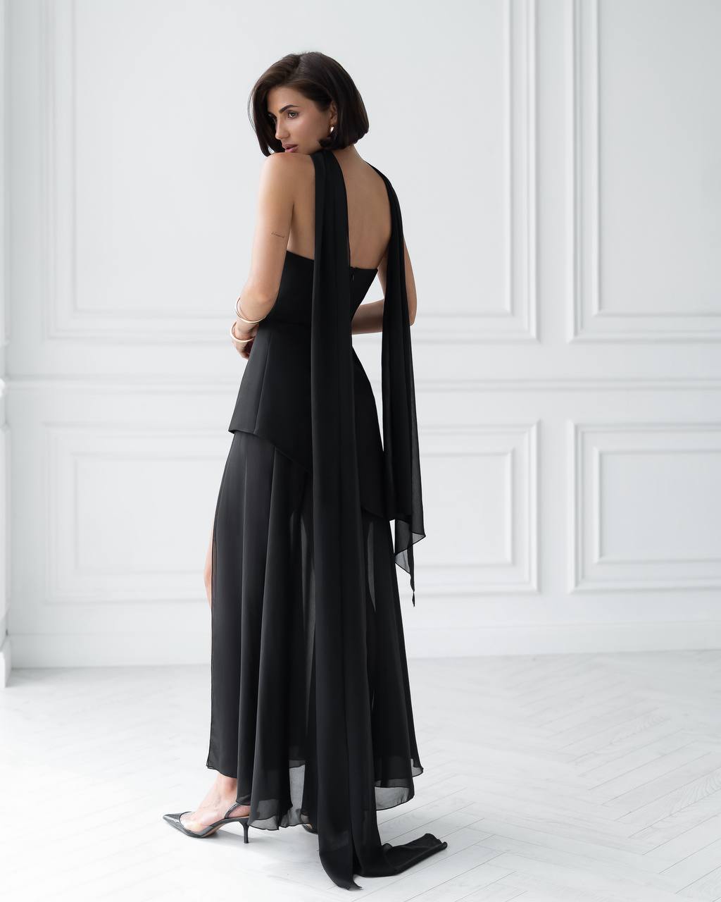 Woman wearing a black dress with a sheer overlay against a white wall.