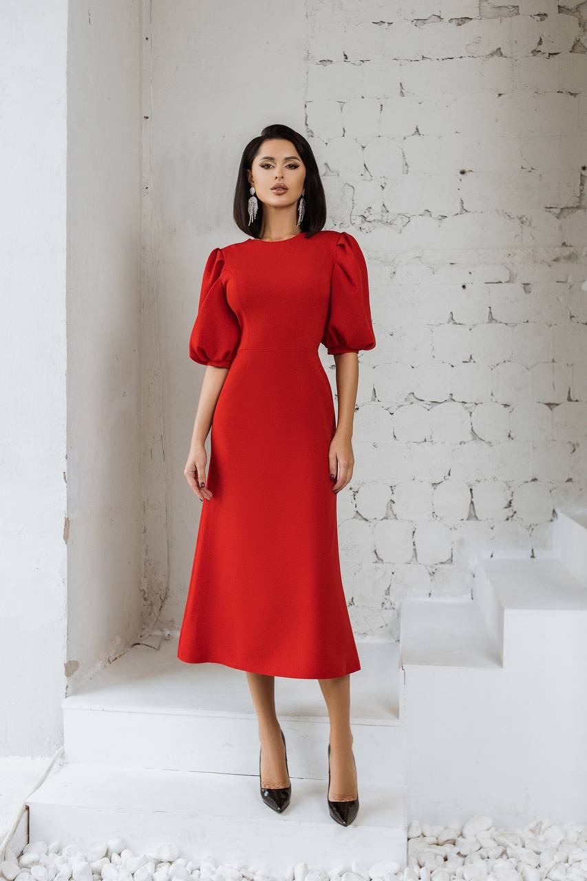 Woman wearing a red dress with puffed sleeves against a white brick wall.