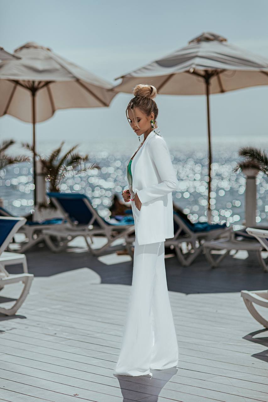 Woman in a white outfit standing on a wooden deck with beach umbrellas and chairs in the background.