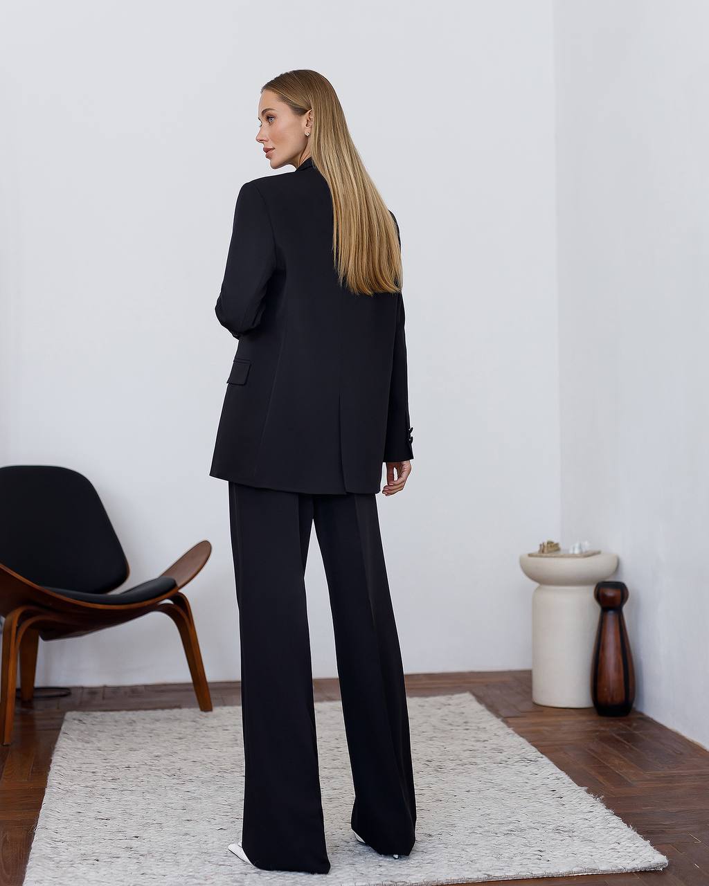 Woman wearing a black suit standing in a minimalistic room with a chair and decorative items.