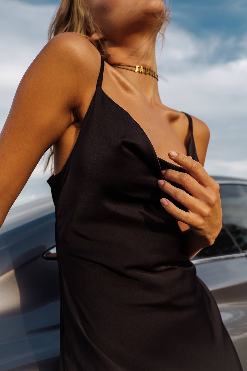 Woman wearing a black dress with thin straps, standing next to a car with a cloudy sky in the background.