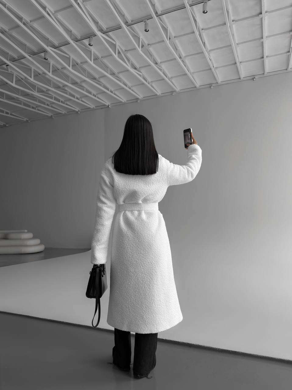 Person in a white coat taking a selfie in a minimalistic room.