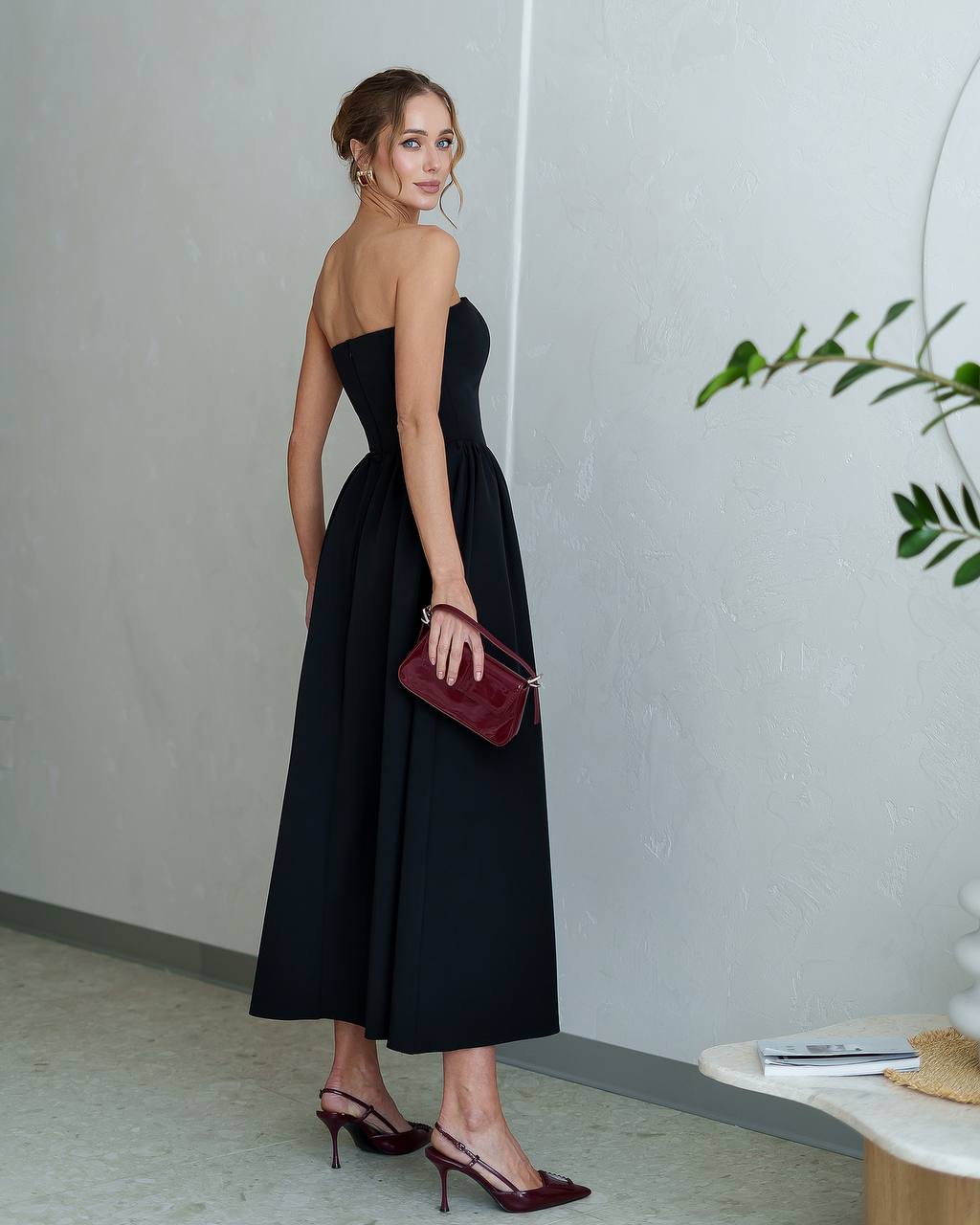 Woman in a black strapless dress holding a red clutch in an indoor setting.