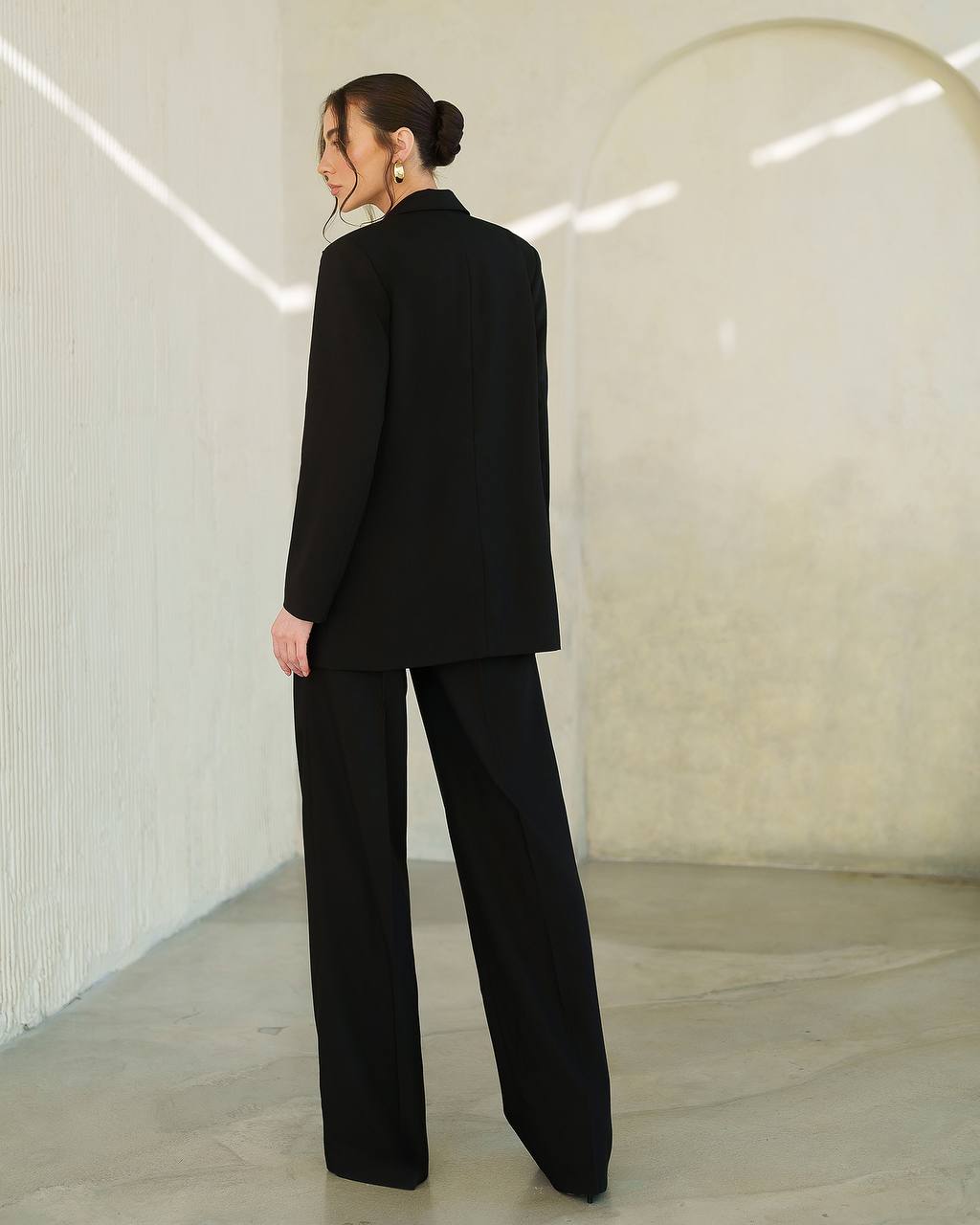 Person wearing a black suit standing in a minimalistic room with white walls and a gray floor.