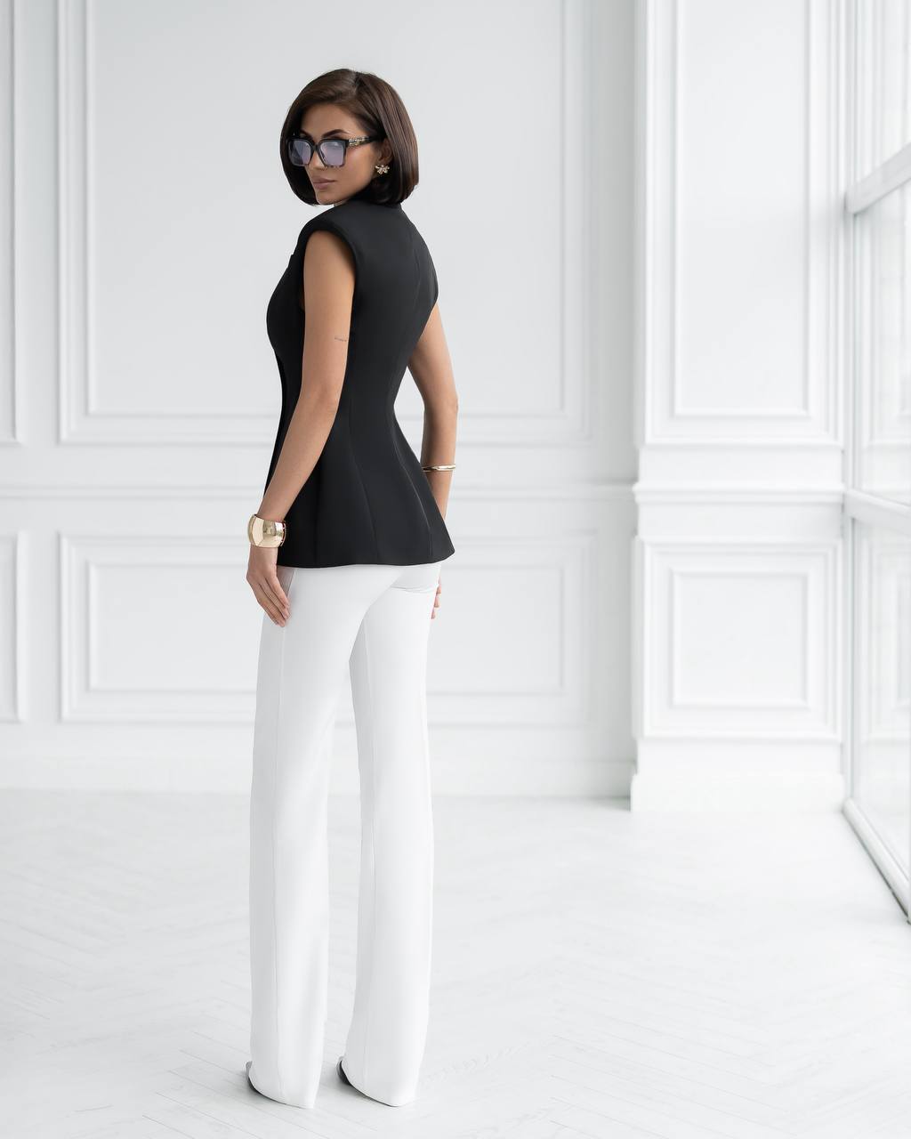 Woman wearing a black sleeveless top and white pants against a white wall.