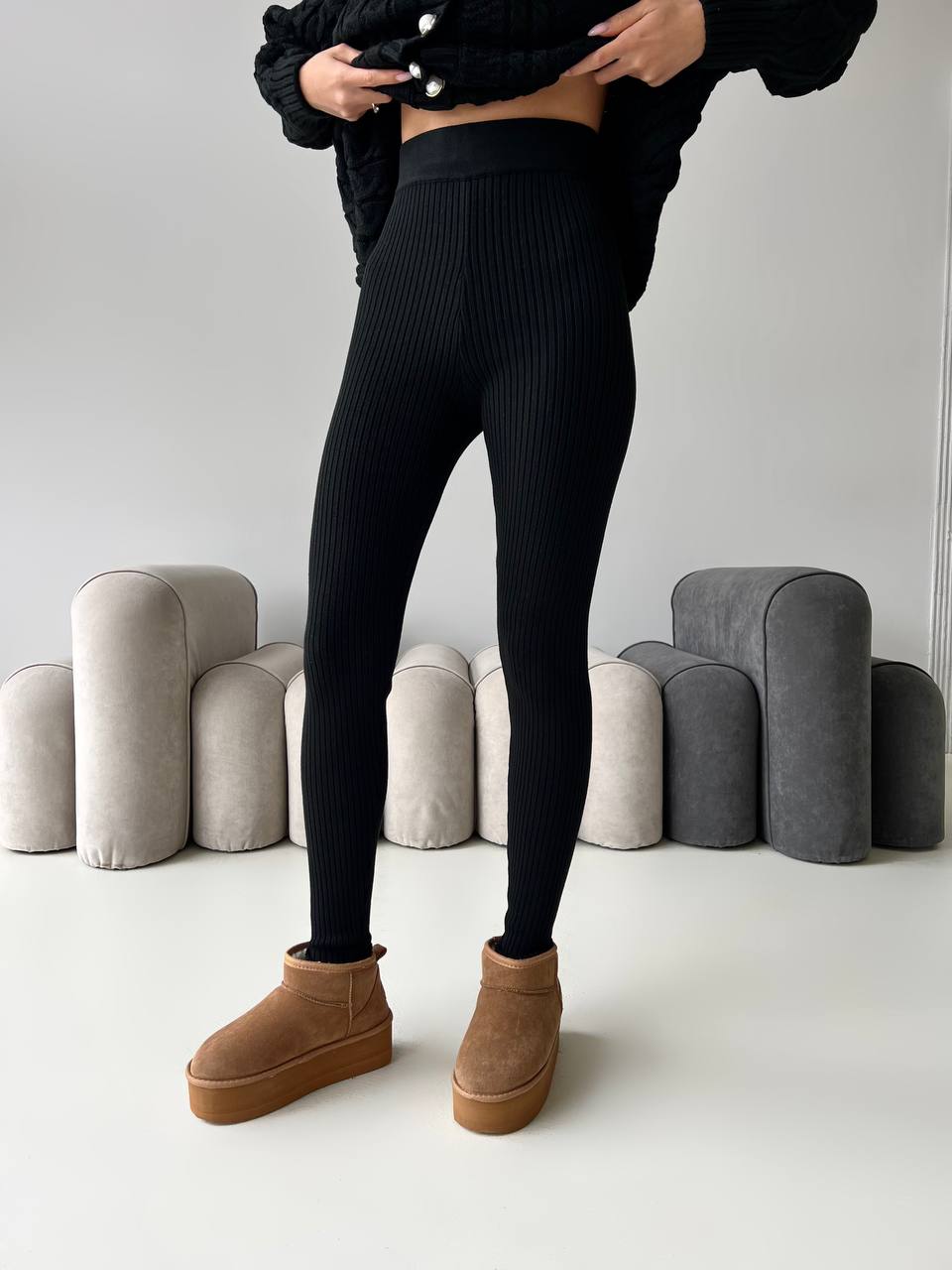 Person wearing black ribbed leggings and brown boots in a minimalistic setting with gray cylindrical objects.