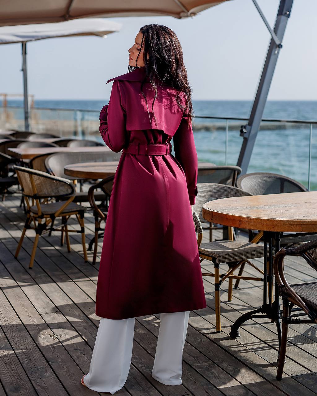 Person wearing a long burgundy coat standing on a wooden deck with tables and chairs, looking out towards the ocean.