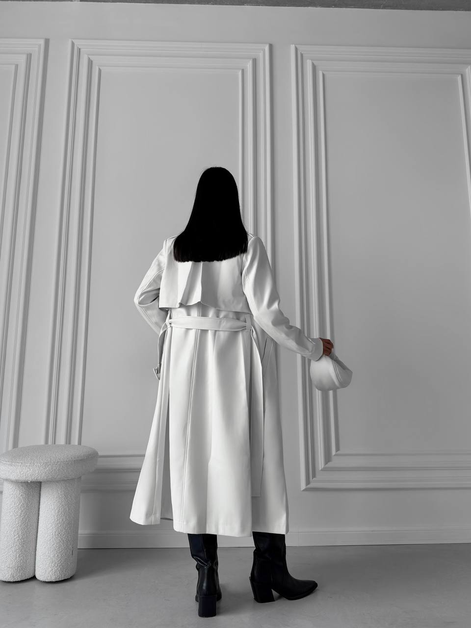 Person wearing a white coat standing against a gray paneled wall.