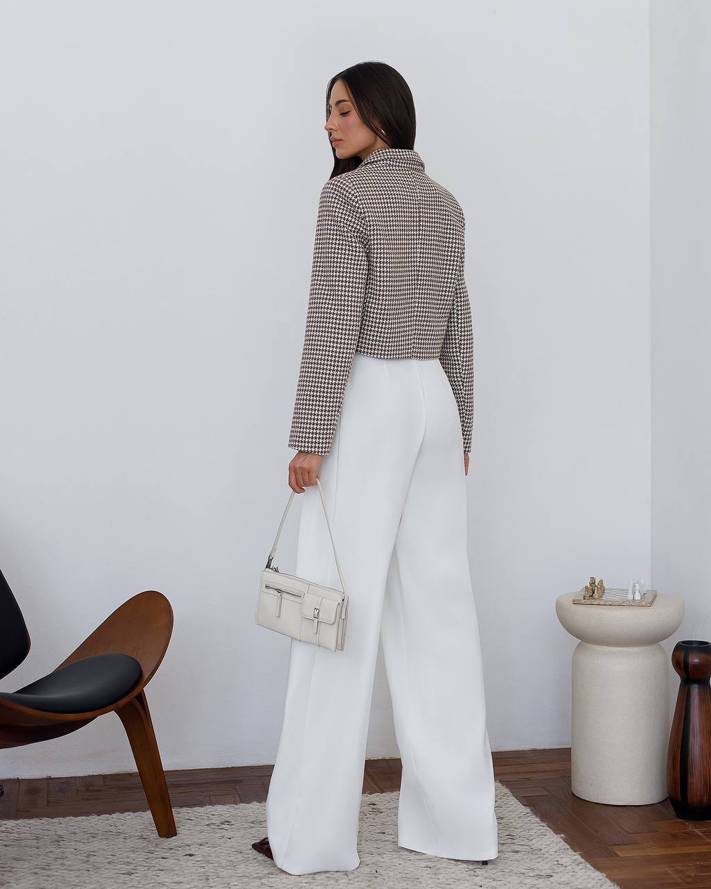 Woman in a checkered shirt and white pants holding a handbag in a minimalistic room.