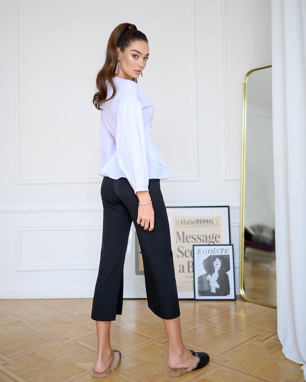 Woman wearing a white blouse and black pants in a room with a mirror and framed picture on the wall.