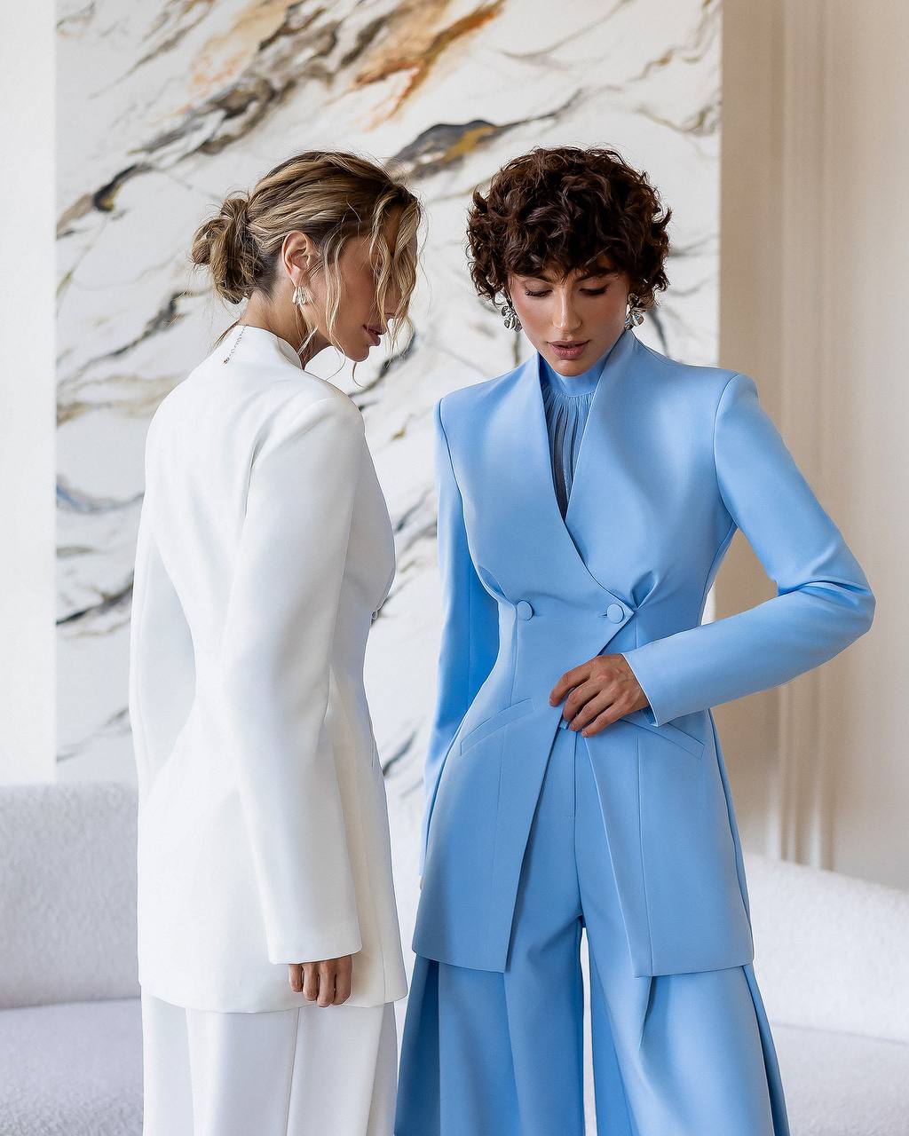 Two women, one in a white coat and the other in a blue suit, standing against a marble wall.