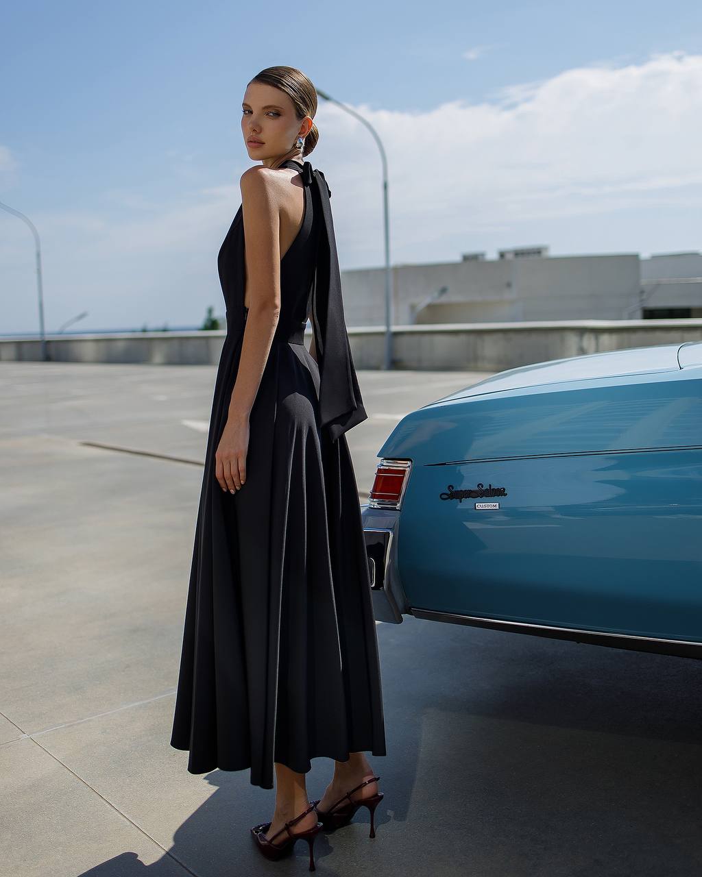 Woman in a black dress standing next to a blue car on a clear day.