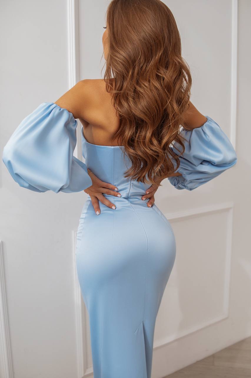 Woman wearing a light blue off-shoulder dress with puffed sleeves against a white wall.