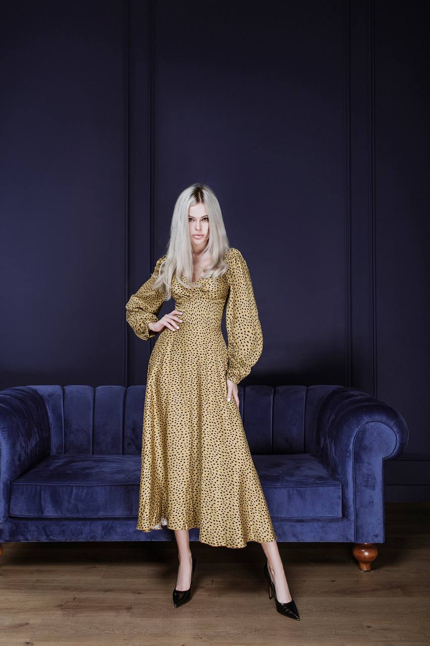 Woman in a gold dress standing in front of a blue couch against a dark purple wall.