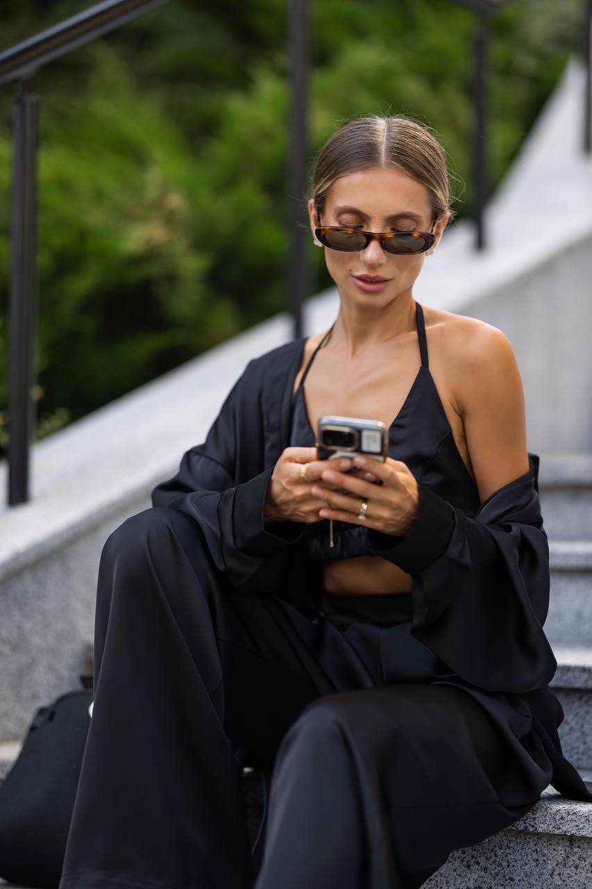 Woman in black outfit sitting on steps using a phone with sunglasses on.