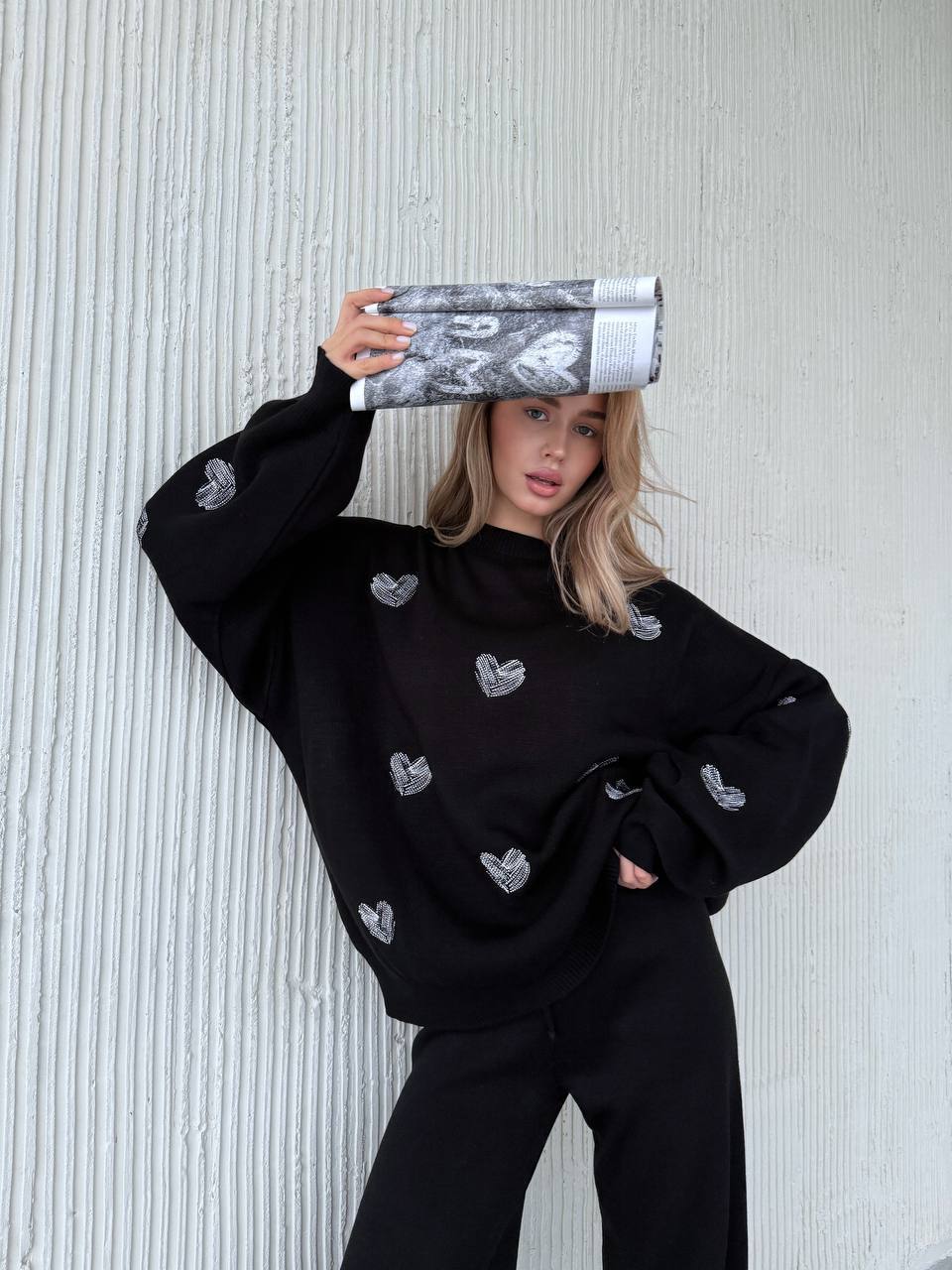 Person wearing a black sweater with heart patterns against a textured white wall.