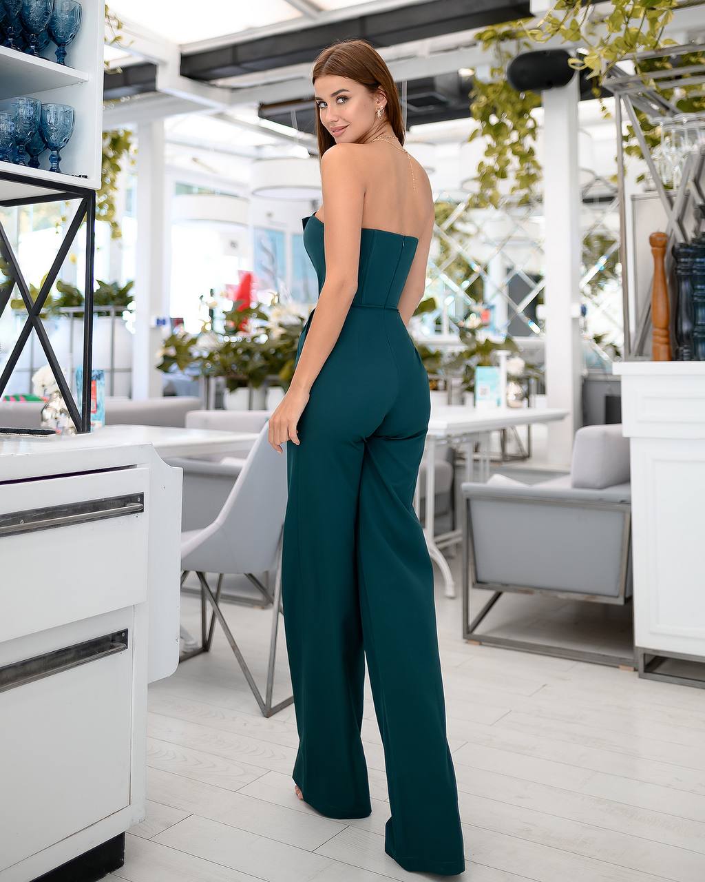 Woman in a teal strapless jumpsuit standing in an indoor setting with white furniture and decor.