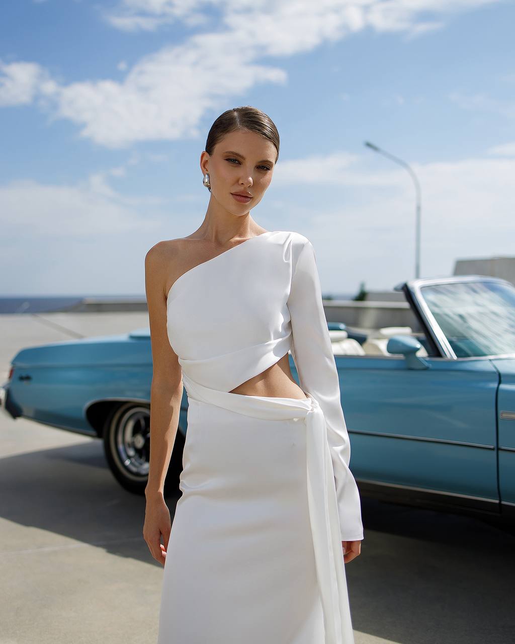 Woman in a white one-shoulder dress standing in front of a blue vintage car.