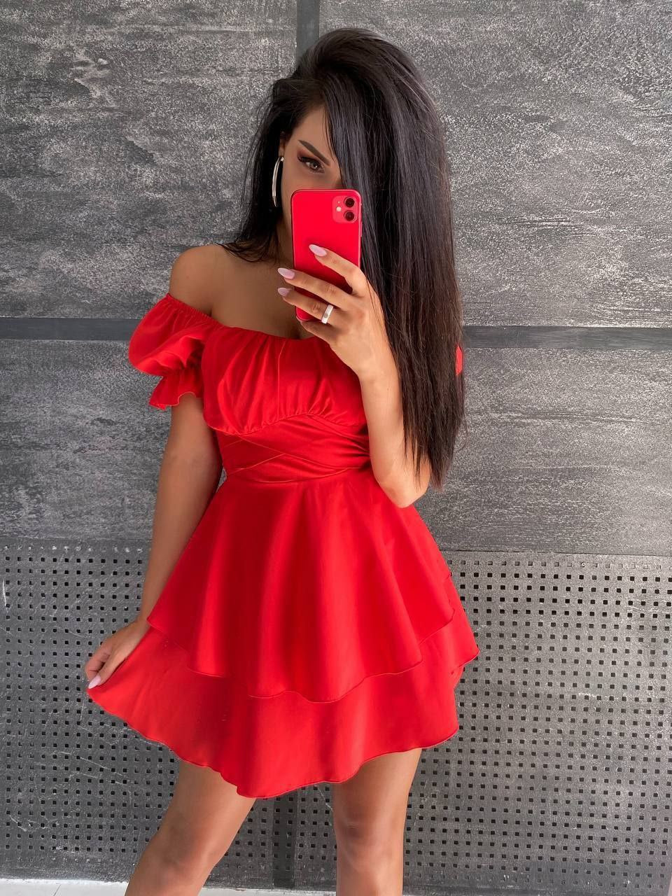 Woman in a red dress taking a mirror selfie against a gray wall.