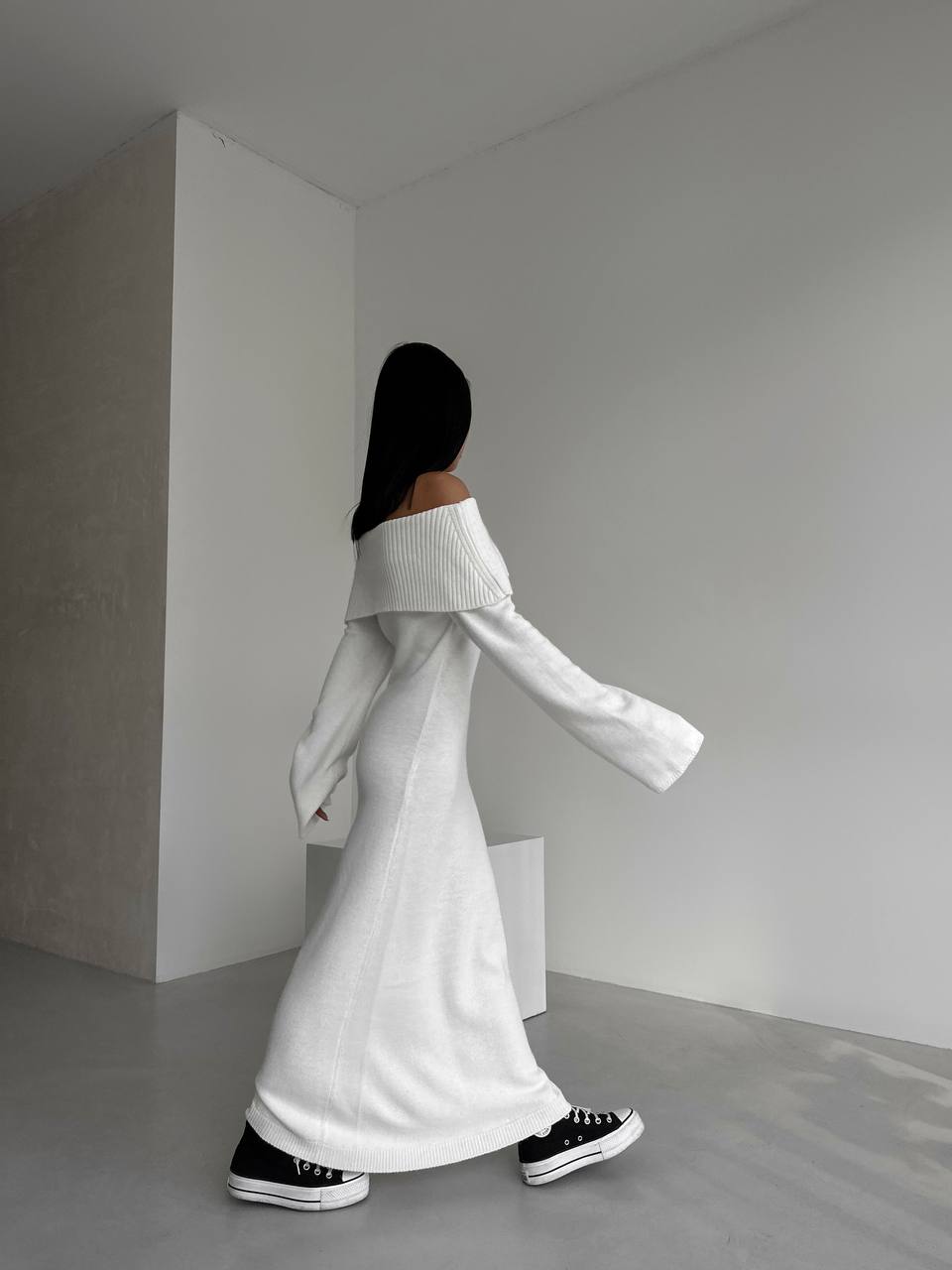 Person wearing a white off-shoulder sweater and black shoes in a minimalistic room.