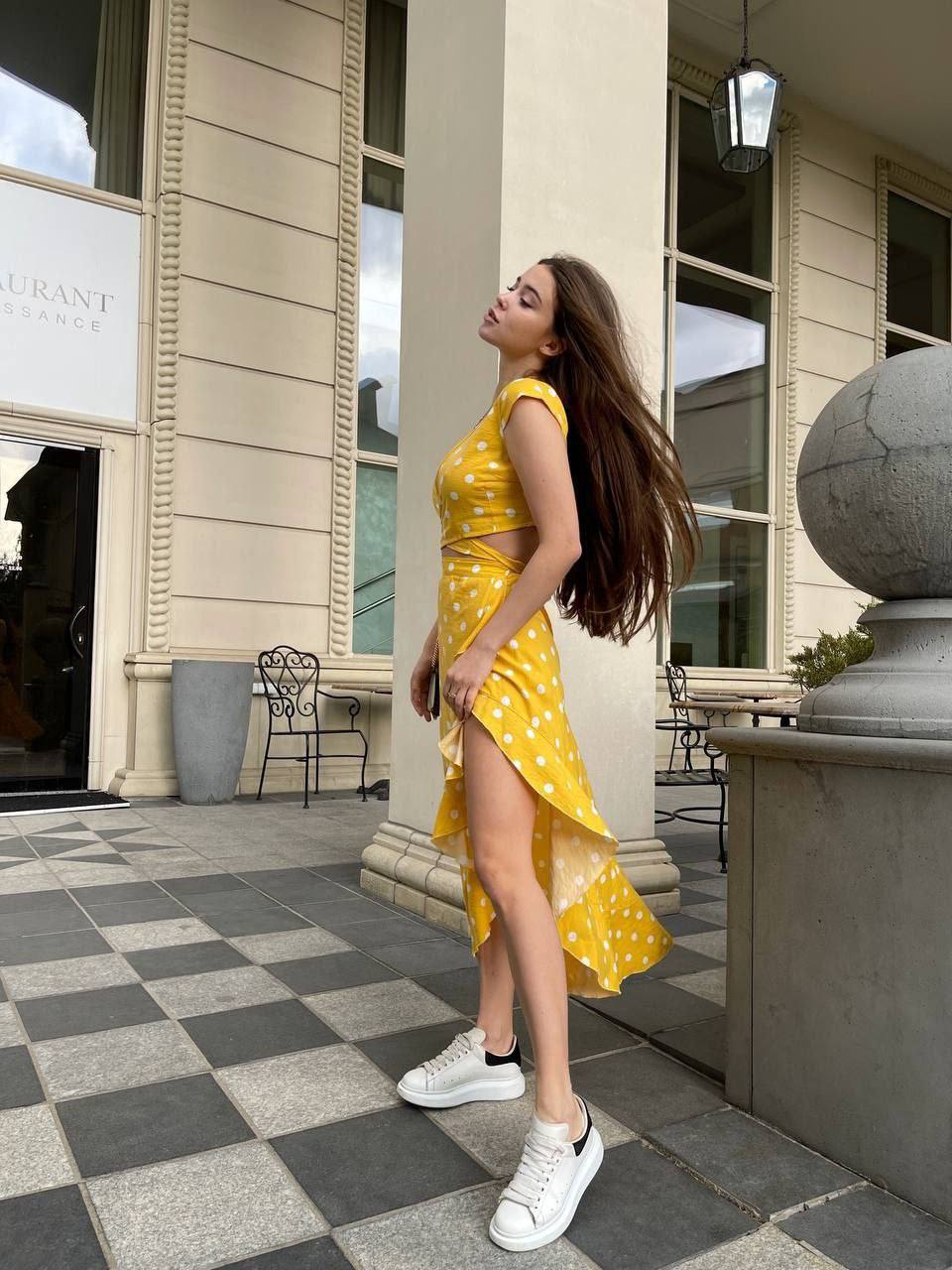 Woman in a yellow dress standing on a checkered pavement outside a building.