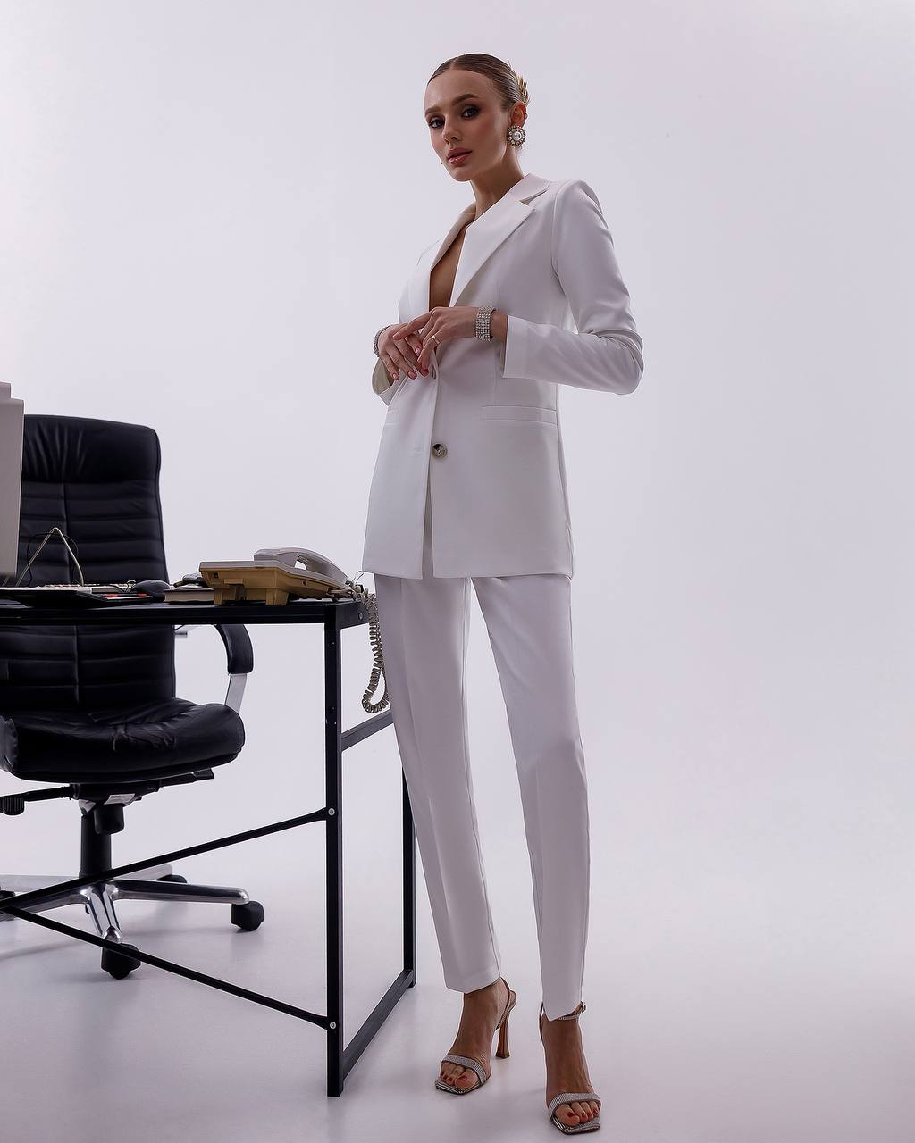 Woman in a white suit standing in an office setting with a desk and chair.