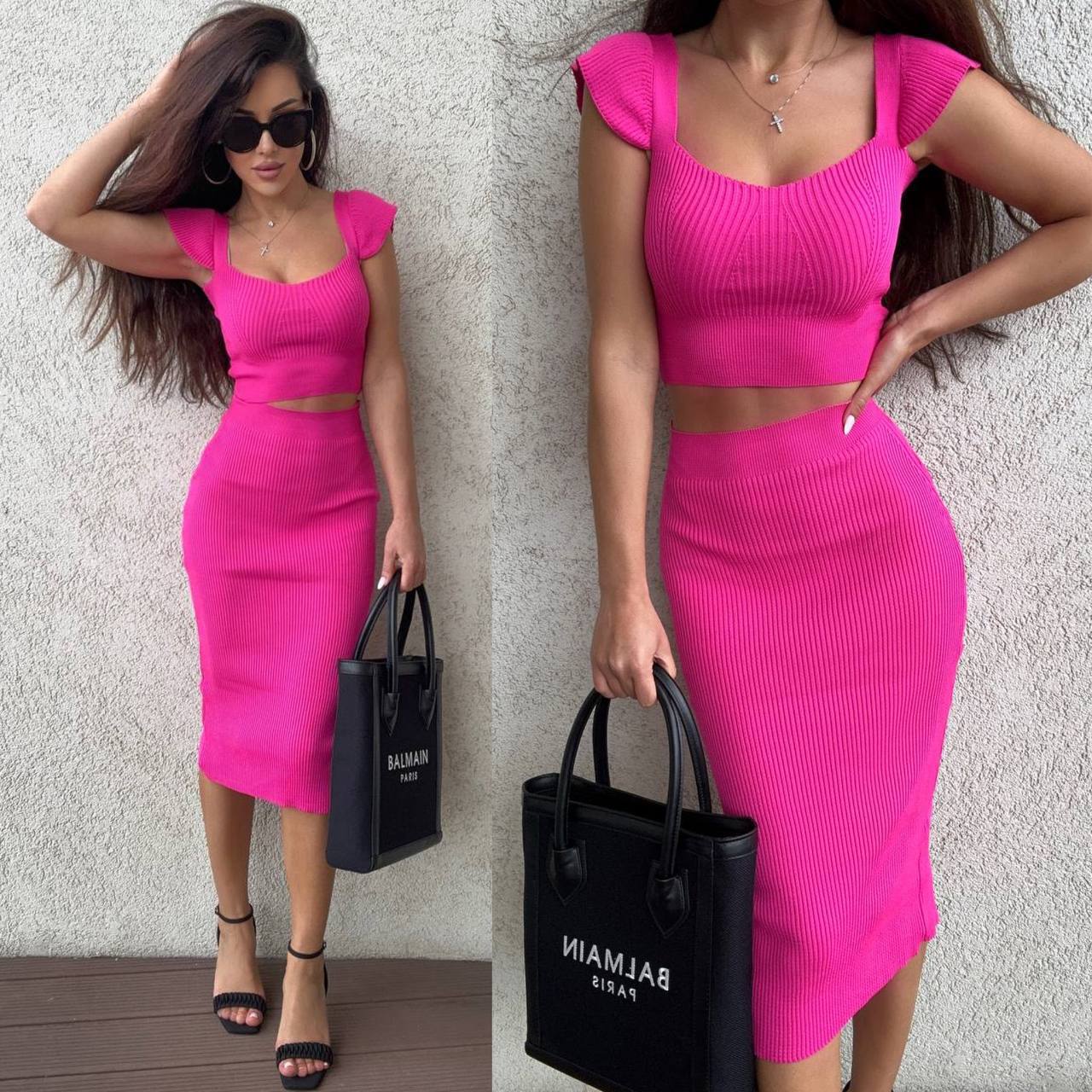 Woman wearing a bright pink two-piece outfit holding a Balmain black handbag against a light gray wall.
