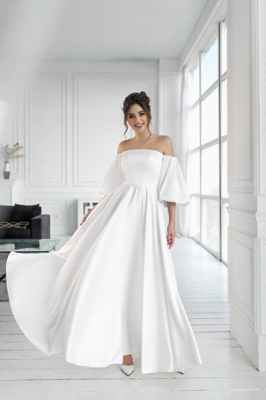 Woman in a white off-shoulder gown standing in a bright room with large windows.