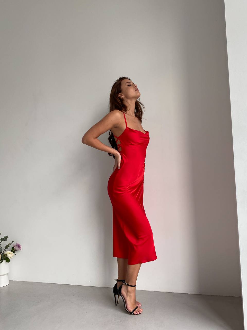 Woman in a red dress standing against a plain white wall with a vase of flowers on the floor.