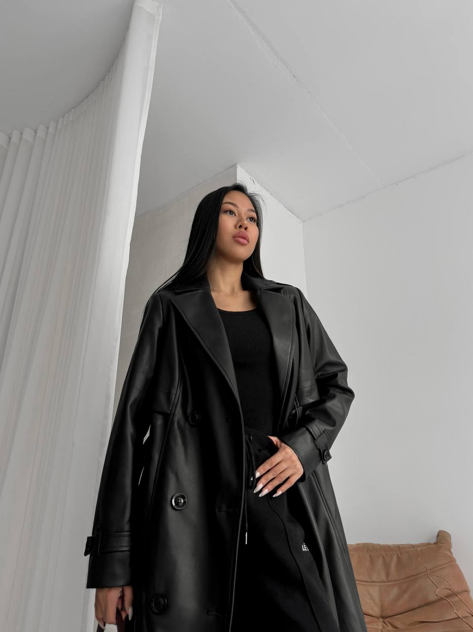 Woman wearing a black coat in a minimalistic room with white walls and a brown couch.