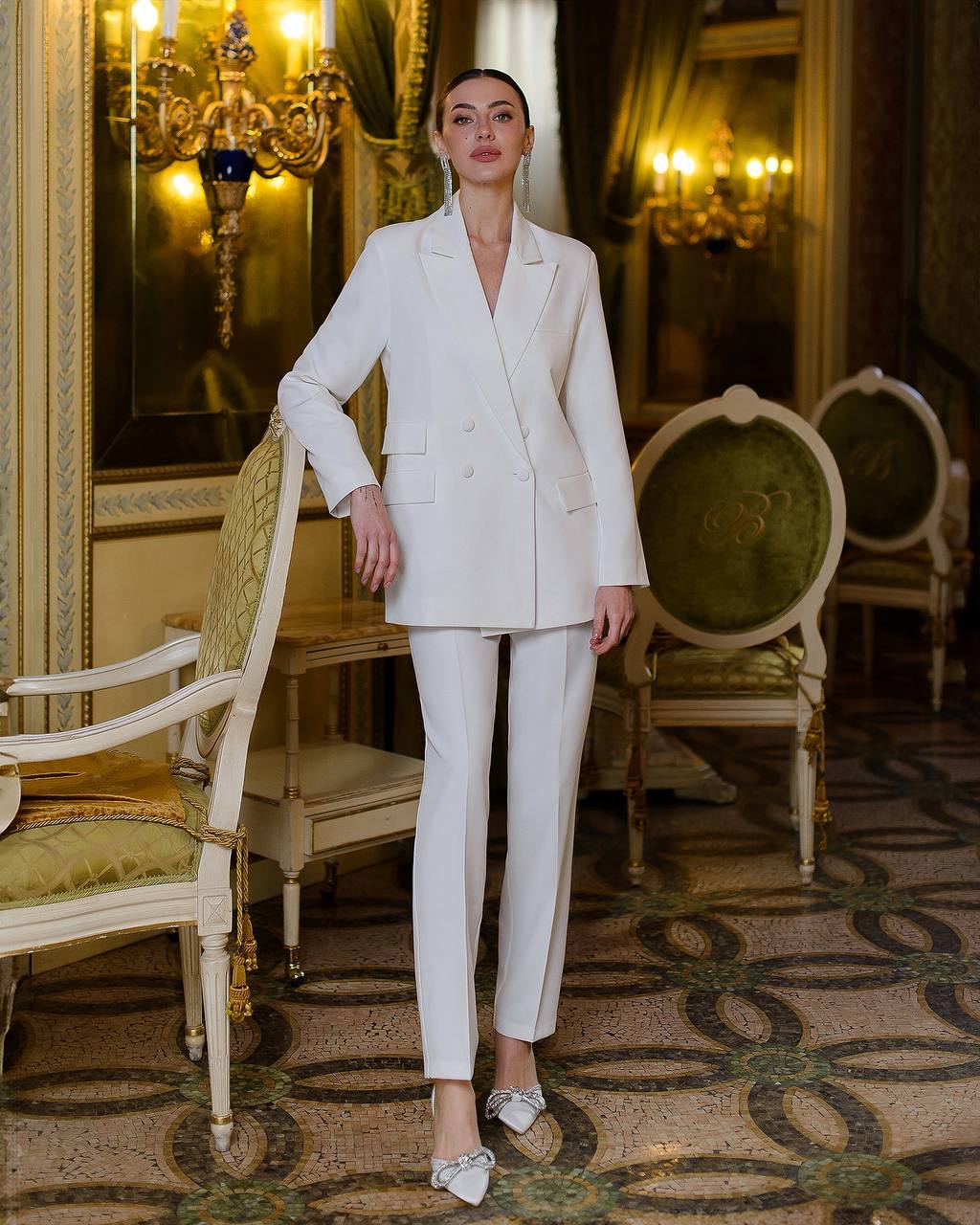Woman in a white suit standing in an ornate room with decorative chairs and mirrors.