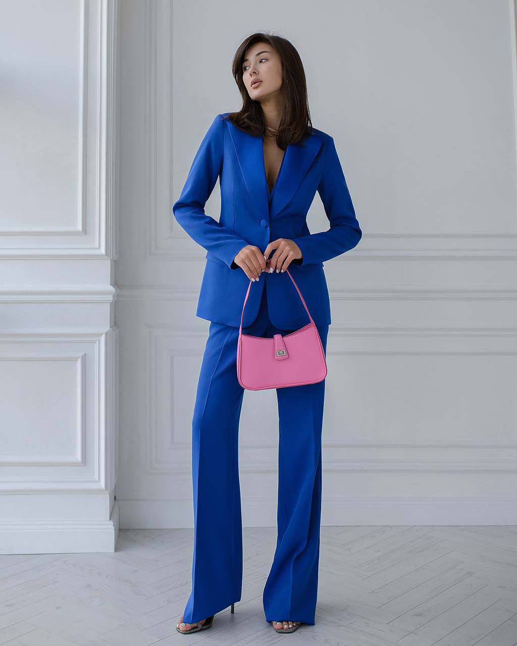 Woman in a blue suit holding a pink handbag against a white wall.