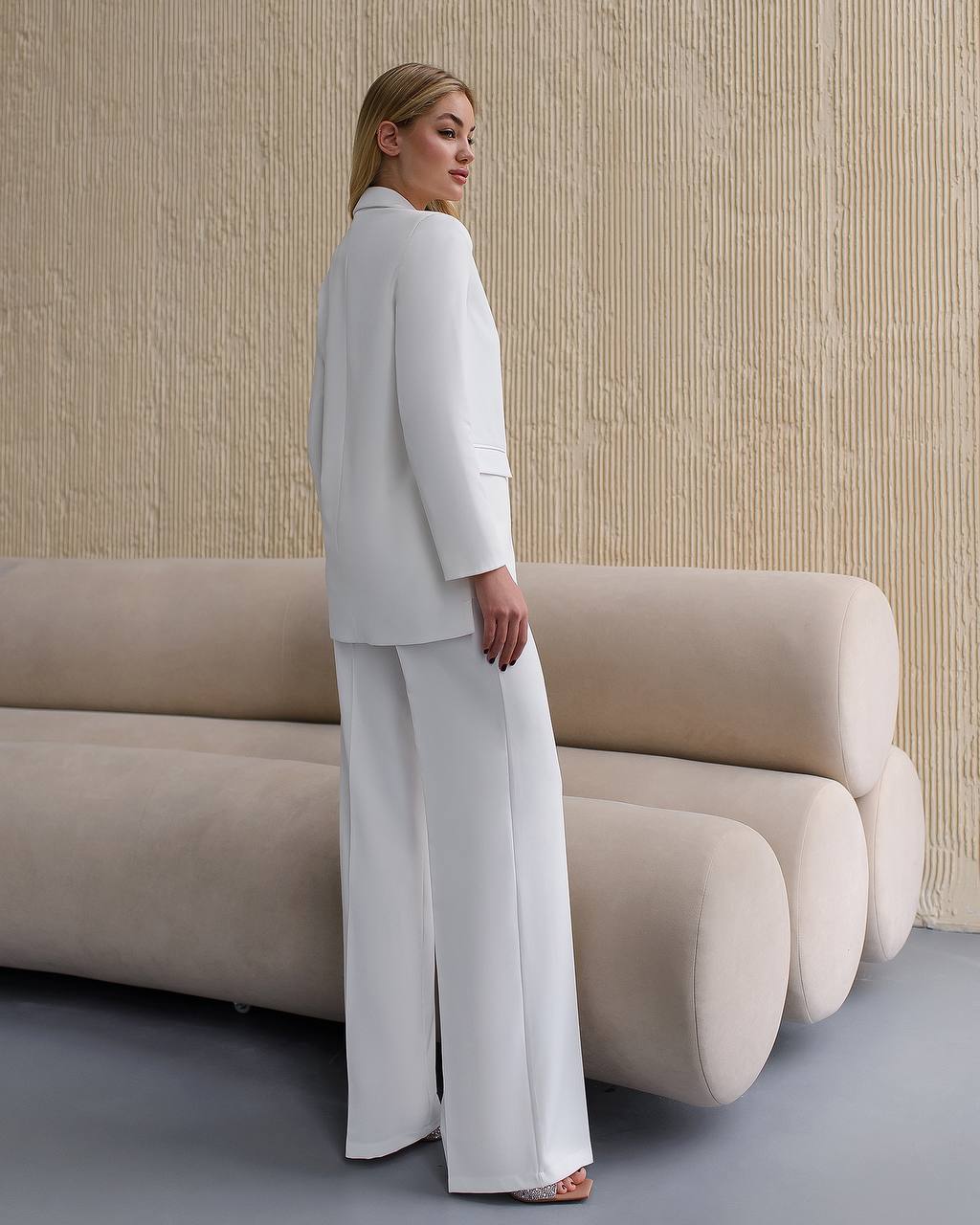 Woman in a white outfit standing next to a modern beige sofa against a textured wall.