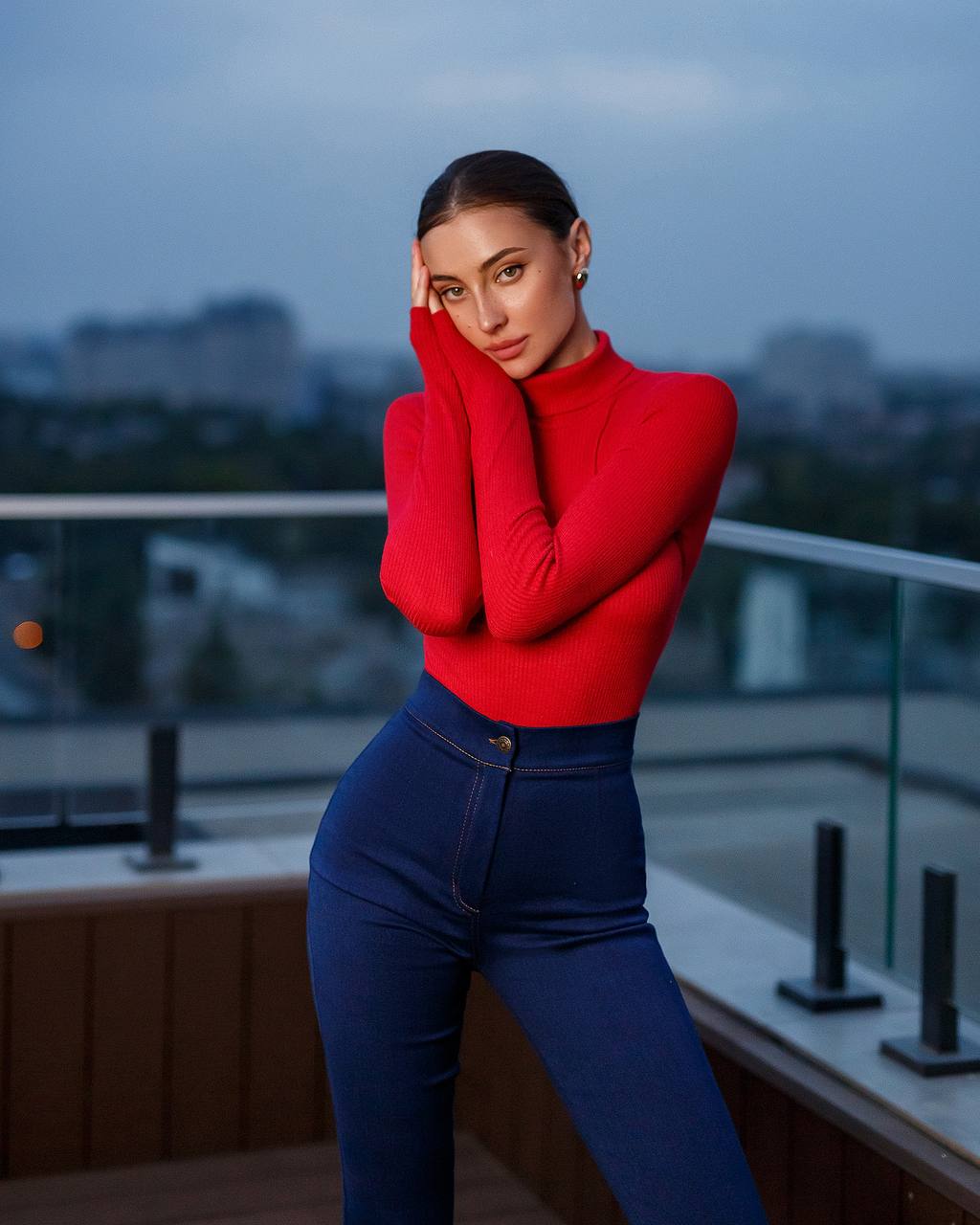 Woman wearing a red sweater and blue jeans standing on a rooftop at dusk.