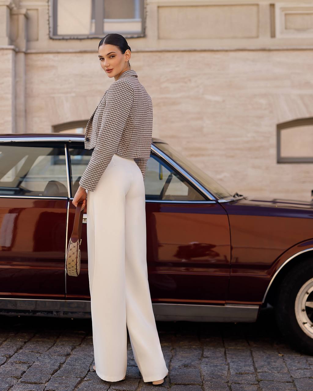 Woman in a patterned blazer and white pants standing next to a vintage car.