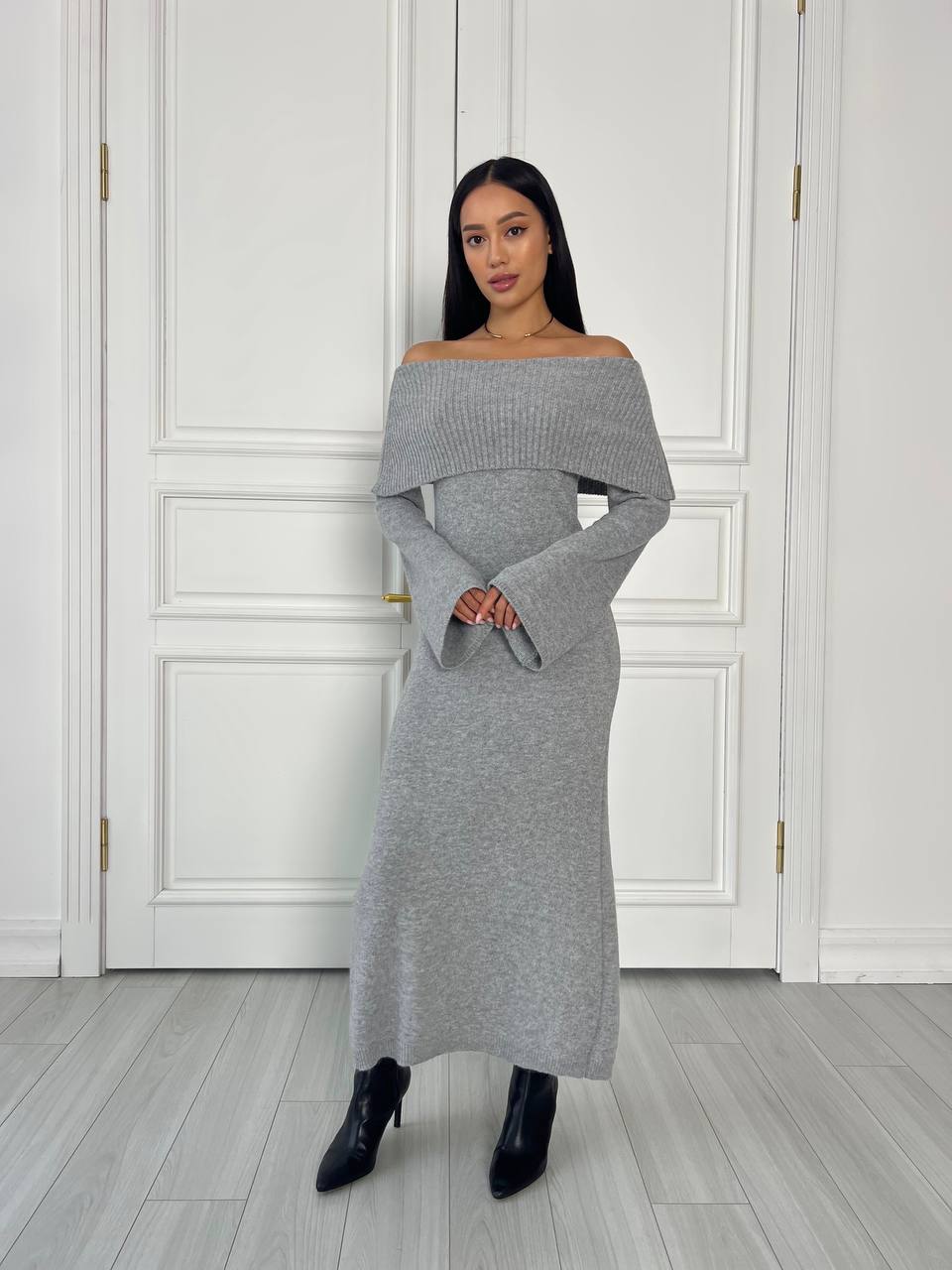 Woman wearing a gray off-shoulder dress standing against a white paneled wall.