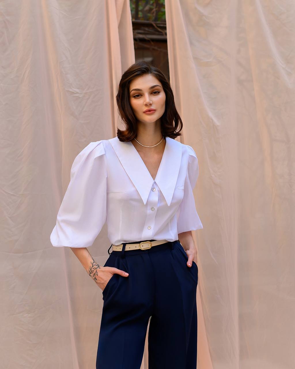 Woman wearing a white blouse and navy pants against a beige curtain background