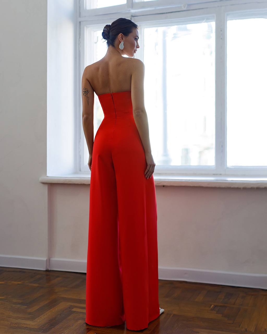 Woman wearing a strapless red jumpsuit standing in a room with large windows.