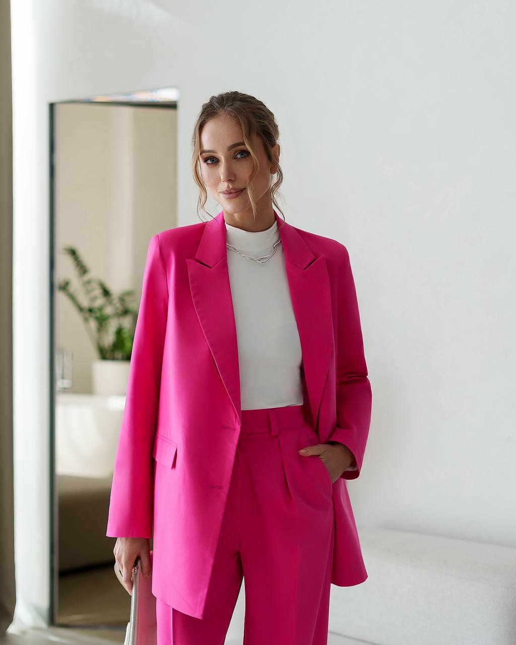 Woman wearing a bright pink suit standing in a room with a mirror and plant.