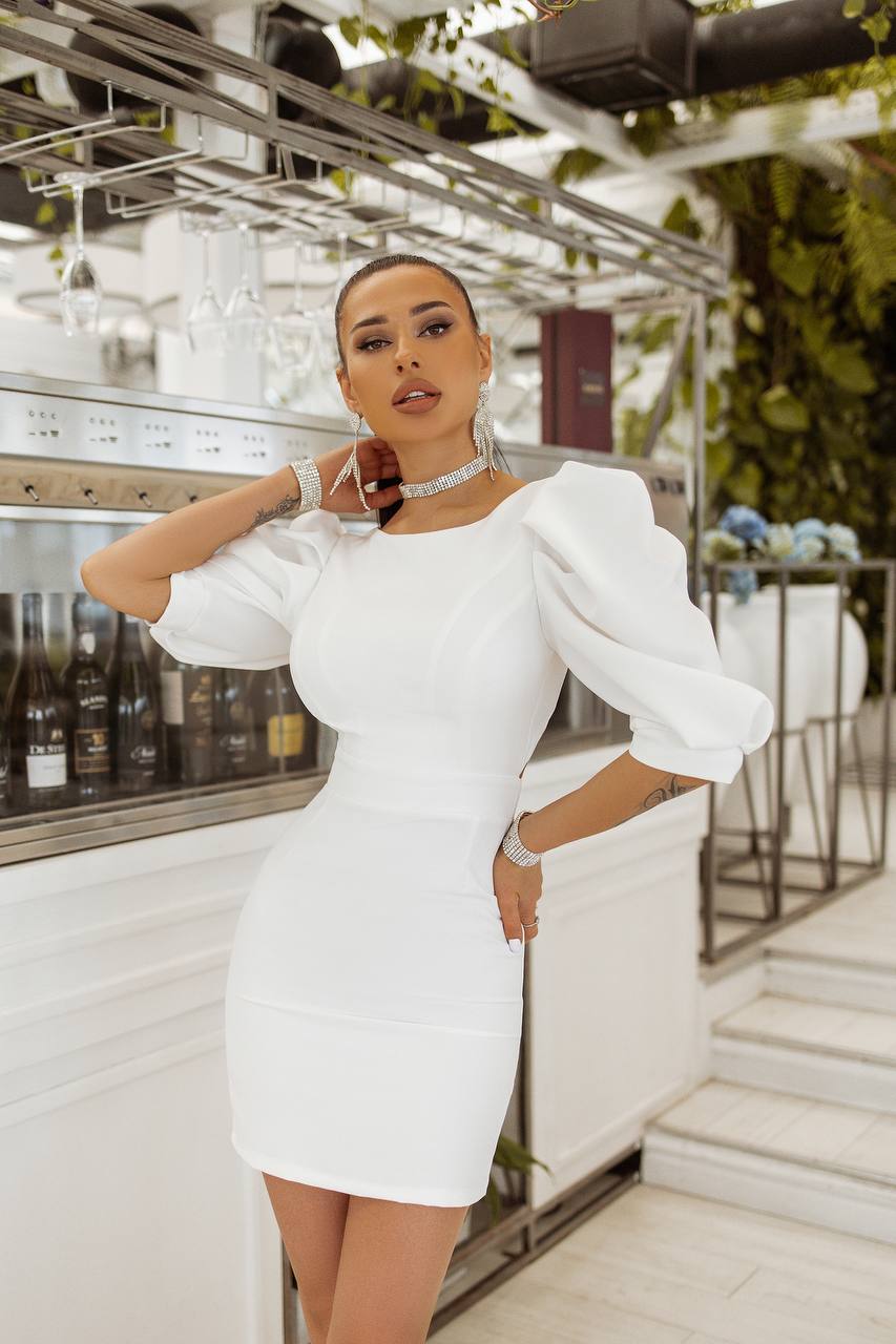 Woman in a white dress standing in an indoor setting with plants and decor.