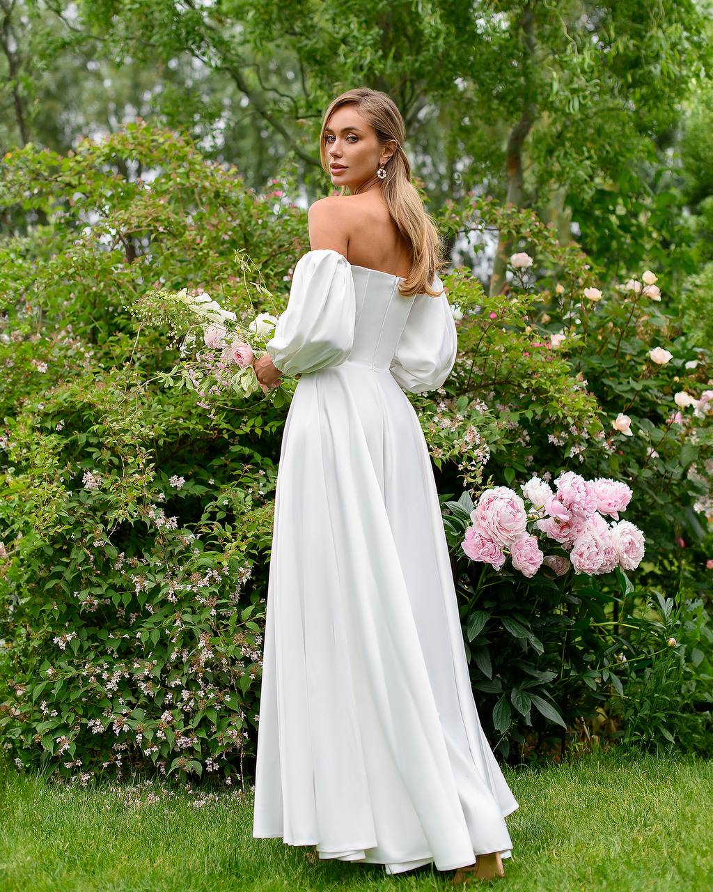 Woman in a white off-shoulder dress standing in a garden with flowers and greenery.