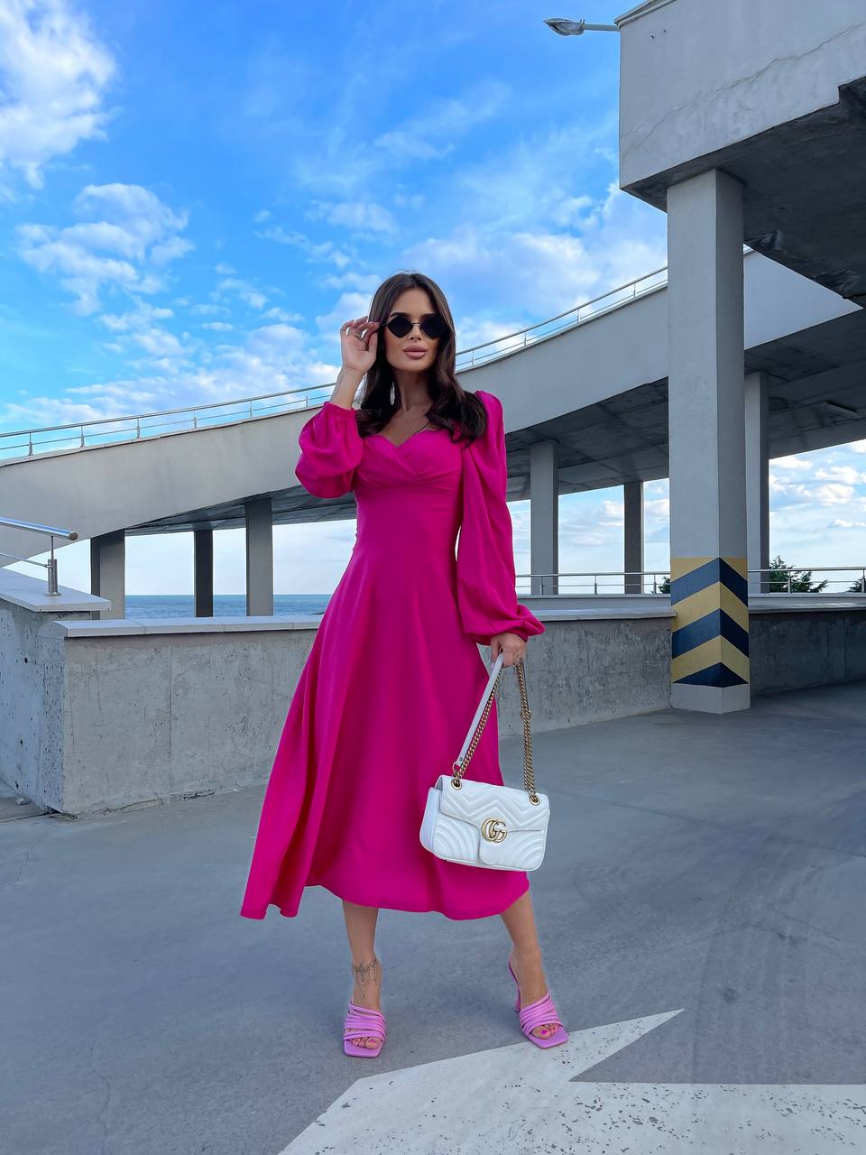 Woman in a bright pink dress holding a white handbag under a blue sky.